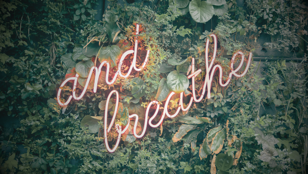 neon letters spelling 'just breath' against a backdrop of green foliage. 