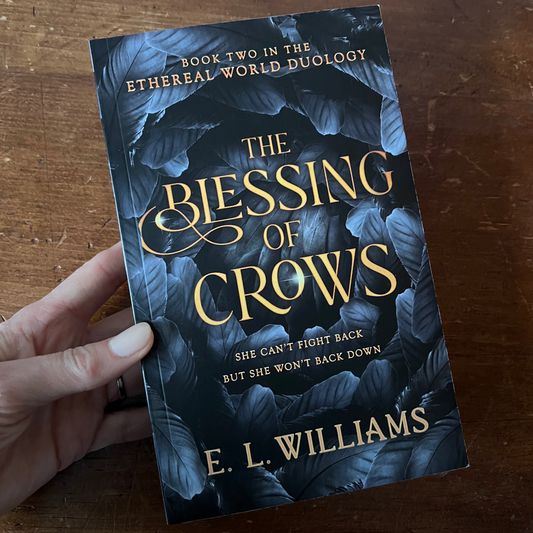 Book titled 'The Blessing of Crows' by E.L. Williams held on a wooden surface